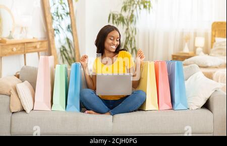 Allegra donna afroamericana con computer portatile e borse per lo shopping acquisto via internet a casa Foto Stock