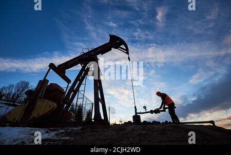 Silhouette di operatore di petrolio in gilet di lavoro e casco usando unità di pompaggio di olio per estrarre petrolio grezzo da pozzo di petrolio, lavorando in campo di petrolio con martinetto a pompa sotto cielo bello. Foto Stock