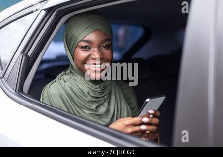 Donna musulmana sorridente nera seduta sul sedile posteriore in auto, utilizzando uno smartphone Foto Stock
