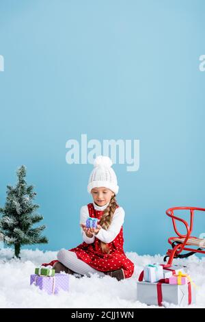 bambino in cappello e vestito invernale seduto sulla neve e. tenuta presente vicino alla slitta isolata in blu Foto Stock