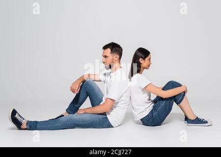 vista laterale di un'elegante coppia interracial in t-shirt bianche, jeans e scarpe da ginnastica che si siedono indietro su bianco Foto Stock