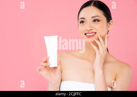 brunette asian donna che tiene tubo di crema cosmetica, viso a contatto e guardando lontano isolato su rosa Foto Stock