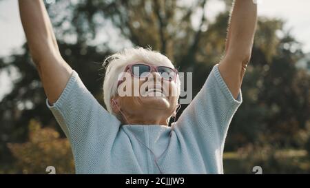Nonna fresca con occhiali da sole rosa e cuffie che si allungano nel parco. Ricreazione nella vecchiaia. Foto di alta qualità Foto Stock