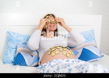 Una donna caucasica incinta si trova sul suo letto e mette le fette di cetrioli sul suo viso e pancia, spa-giorno. Concetto di gravidanza e di cura naturale. Foto Stock