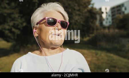 Nonna fresca con occhiali da sole rosa e cuffie che ascoltano musica. Foto di alta qualità Foto Stock