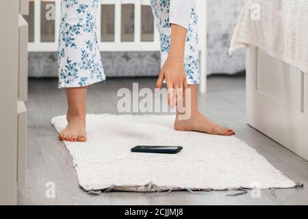 La donna si piega con la mano per prendere il telefono, che è caduto a terra. Primo piano delle gambe. Soggiorno a casa in background. Il concetto di los Foto Stock