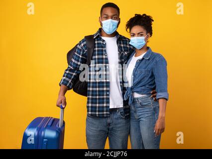 Turisti africani che indossano maschere protettive in piedi con valigia, sfondo giallo Foto Stock