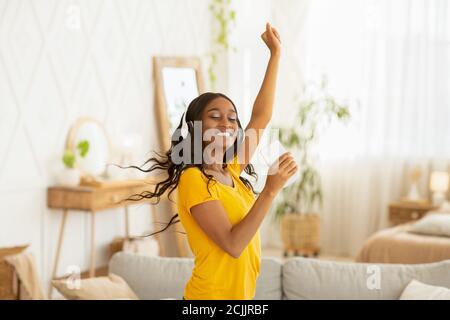 Felice giovane donna nera con cuffie e dispositivo mobile danzare alla sua musica preferita a casa Foto Stock