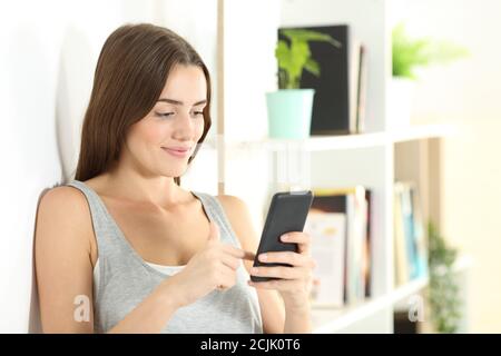 Smiley ragazza che usa il telefono intelligente che controlla il soddisfare in linea che sta in piedi dentro il soggiorno a casa Foto Stock