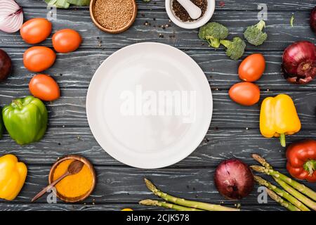 vista dall'alto di verdure fresche e spezie colorate vicino a vuoto piastra su superficie di legno Foto Stock