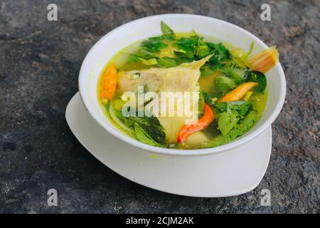 Zuppa di testa di pesce di Gurame o gurame kepala sup ikan con foglie di basilico, pomodori, curry giallo e peperoncino in una ciotola. Cibo indonesiano Foto Stock