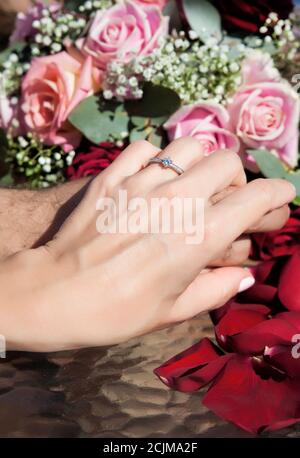 Coppia, donna con anello di fidanzamento diamantato che tiene le mani l'una contro l'altra, set contro un bouquet rosa di rose sullo sfondo e con petali rossi di rose Foto Stock