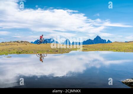 Bella donna che cavalca la sua mountain bike elettrica le tre cime dolomitiche, riflettendosi nelle acque blu di un freddo lago di montagna Foto Stock