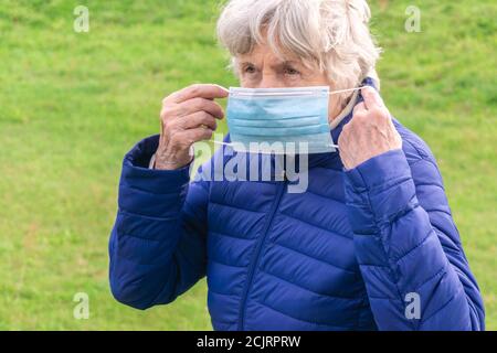 Donna anziana che mette sulla maschera medica nel parco. Donna anziana che toglierà la maschera protettiva all'aperto. Donna anziana con maschera esterna. Protezione da coronavirus Foto Stock