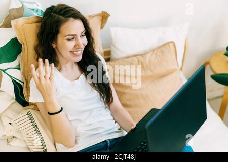 Giovane donna curly affascinante sul letto guarda il portatile e parla. Chat video. Foto Stock