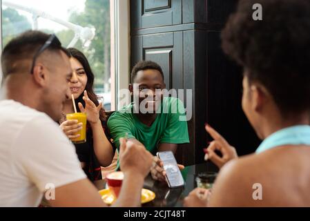 Giovane uomo nero che mostra qualcosa sullo schermo del telefono ai suoi amici mentre si siede in un bar. Foto Stock