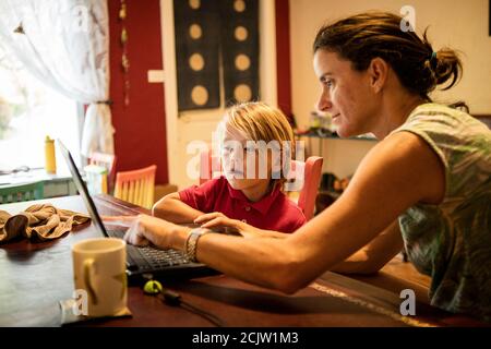 Un ragazzo guarda sopra come sua madre lo aiuta con il lavoro scolastico su un laptop mentre l'apprendimento a distanza ad un tavolo a casa, a Miami, Florida, Stati Uniti Foto Stock