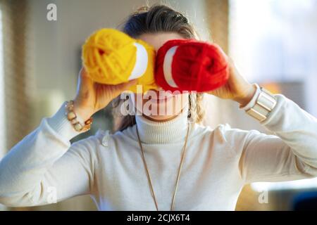 sorridente donna moderna in pullover bianco e gonna fooling intorno con pezzi di filato maglia giallo e rosso nel moderno soggiorno in sole giornate invernali. Foto Stock