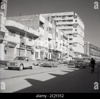 1960, vista da quest'epoca di una strada polverosa e semi-asfaltata a Jeddah, Arabia Saudita, che mostra auto dell'epoca e vari edifici, più vecchi e moderni. Foto Stock