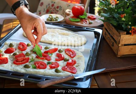 Donna che prepara pane focaccia con ​​tomato a fette, olive e basilico fresco all'aperto. Primo piano. Messa a fuoco selettiva. Cucina italiana. Foto Stock