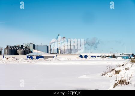 Vista invernale in alcuni luoghi industriali con molti tubi fumosi. Molto freddo giorno di sole, neve copre campo. Grandi costruzioni sono immagazzinate per mulino a vento Foto Stock