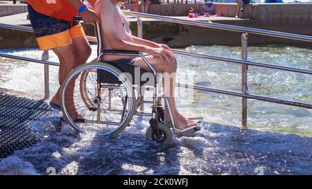 Spiaggia accessibile con sedia a rotelle con rampa per disabili. Uomo su sedia a rotelle che va a nuotare. Foto Stock