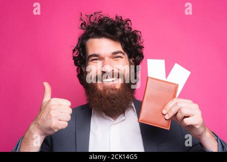 Felice ritratto di bell'uomo bearded in casual mostrando il pollice in su e biglietti di viaggio con passaporto. Foto Stock