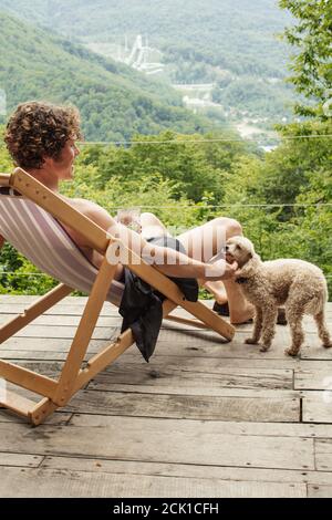 sorridendo un uomo piacevole sta accarezzando un terrier mentre si riposa sul salone. paesaggio di bellezza sullo sfondo della foto. animali, amicizia, tenerne Foto Stock