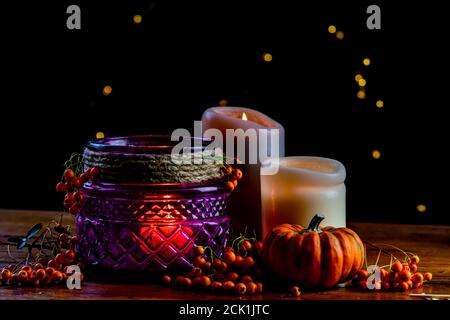 Lanterna viola, zucca dorata arancio bacche rowan e due candele su tavola rustica in legno e sfondo nero con puntini bokeh gialli Foto Stock