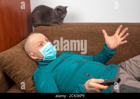 L'uomo in quarantena a casa con una maschera medica sul viso si trova sul divano e guarda la TV accanto a un gatto grigio. Riposo durante l'epidemia di coronavirus. Foto Stock