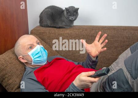 L'uomo in quarantena a casa con una maschera medica sul viso si trova sul divano e guarda la TV accanto a un gatto grigio. Riposo durante l'epidemia di coronavirus. Foto Stock