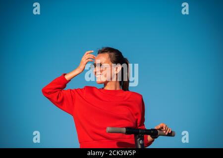 Bella donna sorridente in rosso elegante top e occhiali da sole isolati su sfondo blu cielo tenendo il manubrio di scooter elettrico e guardando avanti. Foto Stock