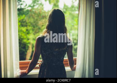 Una giovane donna si sta alzando al mattino e. sta aprendo le tende per guardare all'esterno Foto Stock