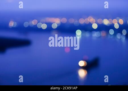 Defocalizzare il paesaggio urbano su un fiume nella notte blu, luminoso e colorato sfocato con bokeh cerchio intorno luci colorate, nave container su un fiume. Foto Stock