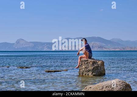 L'uomo asiatico anziano in maschera di faccia siede da solo su un pietra in una baia di mare Foto Stock