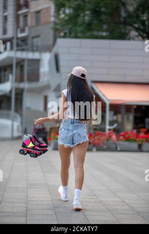 Ragazza dai capelli scuri in pantaloncini e top con pattini a rotelle in mani Foto Stock