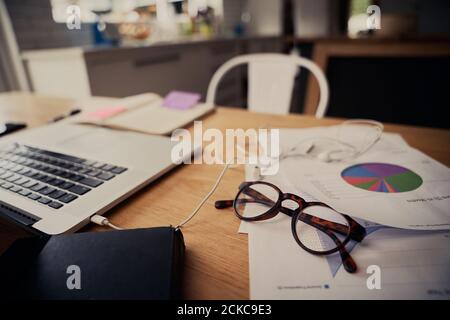 Closeup di occhiali sui documenti vicino al computer portatile con gli auricolari collegati su una scrivania in legno a casa Foto Stock