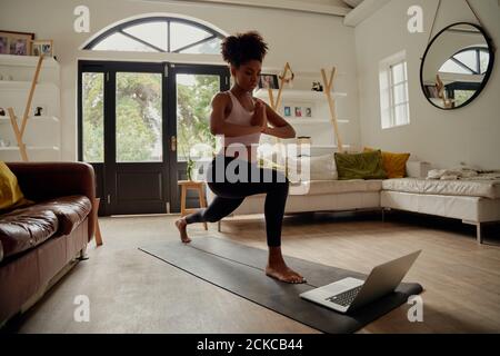 Donna africana sana guardando video sul computer portatile mentre si esercita con mani unite sul tappetino yoga a casa Foto Stock