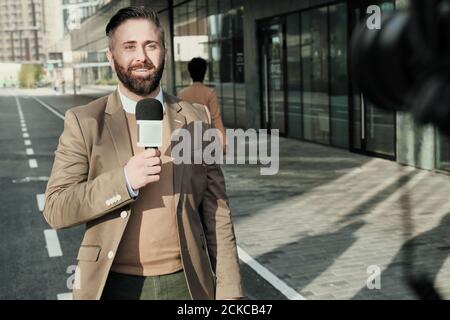 Uomo maturo che parla in microfono e sorridente alla telecamera durante la trasmissione in diretta sulla strada cittadina Foto Stock