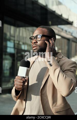 Giornalista africano in occhiali che parla in microfono durante la trasmissione dal vivo in città Foto Stock