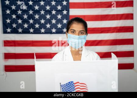 Dottore o infermiera che viene alla cabina di polling con maschera medica che indossa per il voto - concetto di elezione degli Stati Uniti, in persona voto mostrando con bandiera degli Stati Uniti come Foto Stock