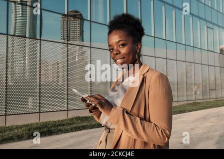 Ritratto di giovane donna africana che sorride alla macchina fotografica durante la digitazione un messaggio sul suo cellulare in città Foto Stock