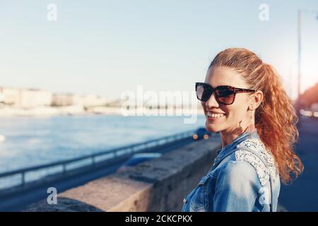 Giovane donna rossa in occhiali da sole con un sorriso toothy in città ritratto sulla riva del fiume Foto Stock