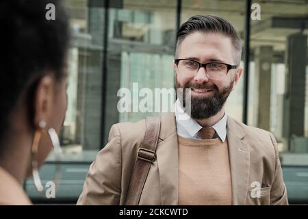Uomo d'affari bearded in occhiali sorridente durante la conversazione con il suo collega in città Foto Stock