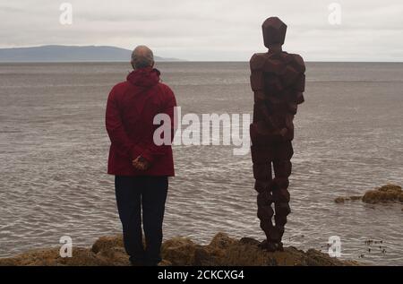 La scultura, IL GRIP di Anthony Gormley e un uomo 60+ in piedi su rocce a Saddell Bay, Kintyre, Argyll, Scozia, guardando insieme Kilbrannan Sound Foto Stock