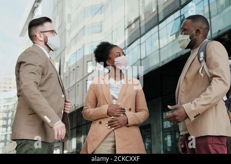 Gruppo di colleghi che indossano maschere di protezione con cui parlano l'uno con l'altro mentre si trova all'aperto vicino all'edificio dell'ufficio Foto Stock