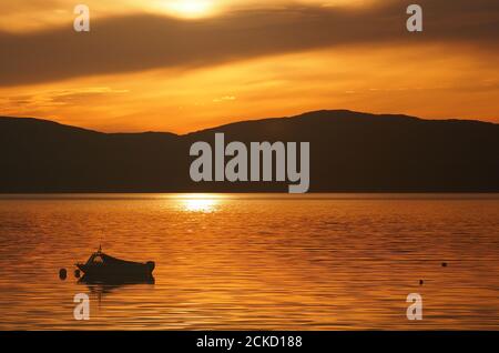Tramonto a Carsaig Bay, Tayvallich, Argyll, Scozia con la silhouette di una piccola barca a motore in primo piano e che si affaccia sul Sound of Jura a Jura Foto Stock