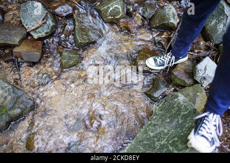 Un giovane uomo con pantaloni e sneaker blu si arrampica attraverso un ruscello che scorre su pietre colorate. Luce naturale con spazio per la copia. Foto Stock