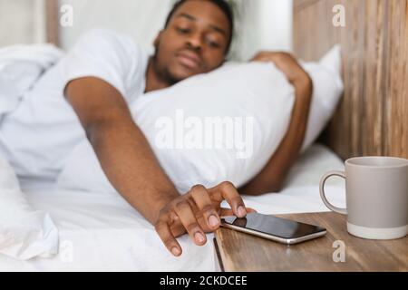 Sleepy Black Man Reaching per smartphone disattivazione sveglia Indoor Foto Stock