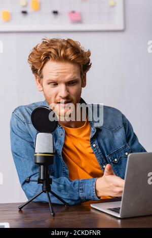 giovane emittente in camicia denim che punta con la mano verso il computer portatile mentre si parla nel microfono Foto Stock